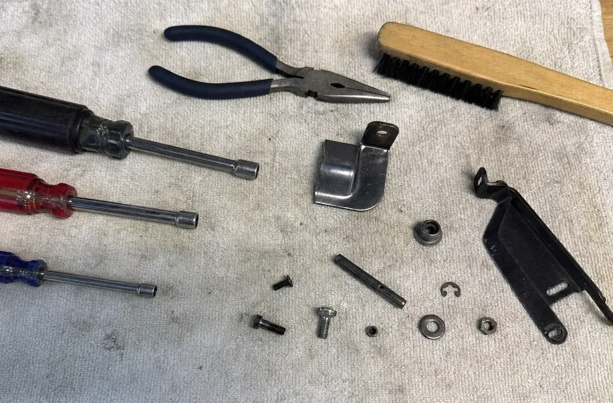 Pliers, a wooden-handled brush and nut drivers on a canvas cloth beside a bracket, pins and small screws